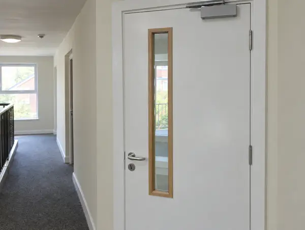 White timber door with a narrow vertical glass vision panel in a flats corridor